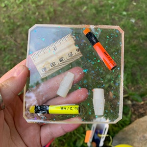 May include: A clear resin tray with a ruler, two pencil erasers, and two pencils. The pencils are labeled "10 1/2 HB" and "2 HB". The tray has a glittery, iridescent finish.