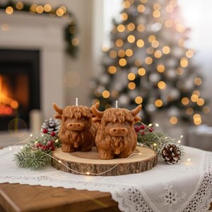 May include: Two brown Highland cow candles with white wicks sit on a wooden slice, decorated with pinecones and greenery. The candles are on a table with a white lace tablecloth, in front of a fireplace and Christmas tree.