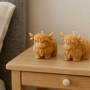 May include: Two tan-colored Highland cow candles with small horns and shaggy fur are displayed on a wooden nightstand. The candles have a detailed design and a small white wick. The background includes a bed and a wall.