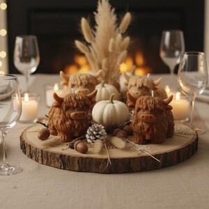 May include: A beautifully set dining table with white plates, gold cutlery, and wine glasses. The centerpiece features Highland cow candles, a white pumpkin, and decorative elements on a wooden slice, creating a cozy, autumnal atmosphere.