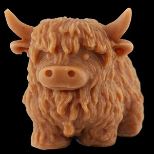 May include: A light brown, sculpted figurine of a Highland cow. The cow has a shaggy coat, small horns, and a detailed face. The figurine is set against a black background.