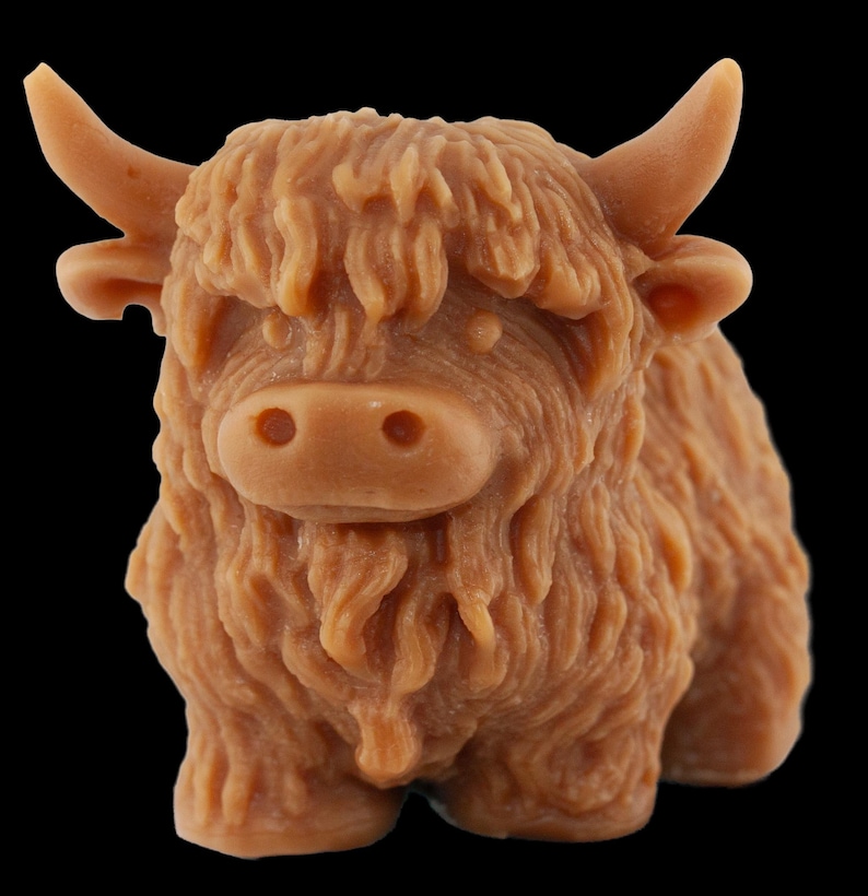 May include: A light brown, sculpted figurine of a Highland cow. The cow has a shaggy coat, small horns, and a detailed face. The figurine is set against a black background.