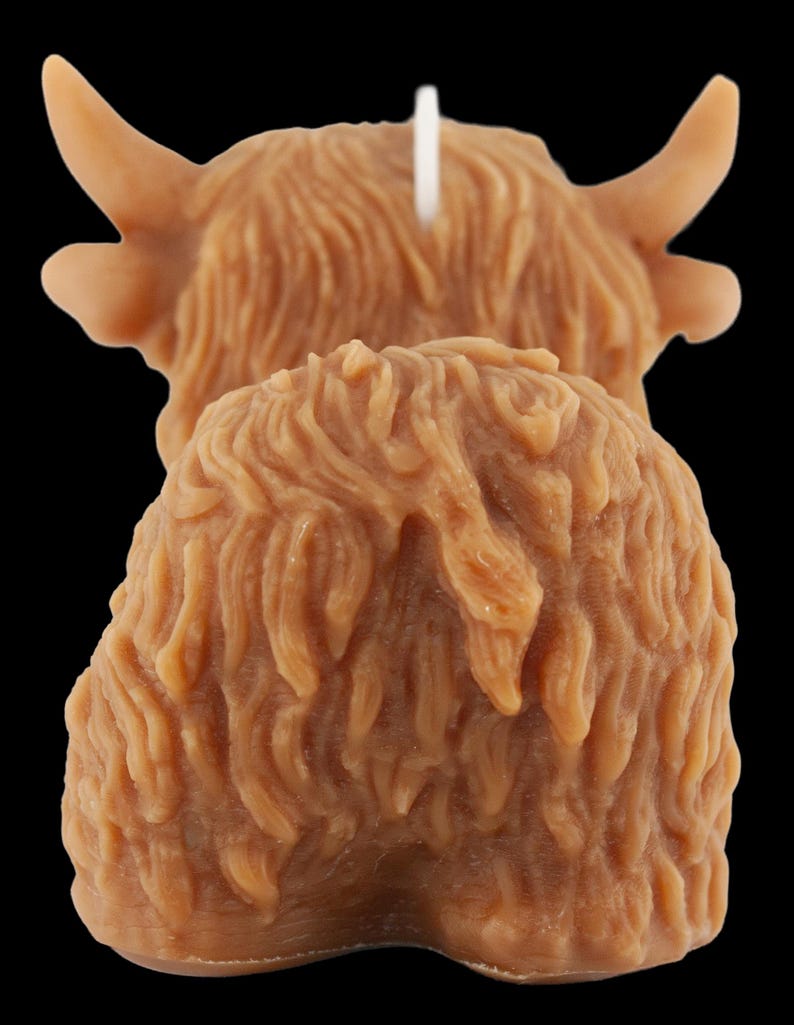 May include: A brown candle shaped like a Highland cow, featuring detailed fur texture and curved horns. A white wick is visible. The candle is set against a black background.