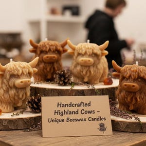 May include: Handcrafted Highland cow beeswax candles in various shades of brown and tan. The candles are shaped like Highland cows and are displayed on wooden slices. The sign reads "Handcrafted Highland Cows - Unique Beeswax Candles."