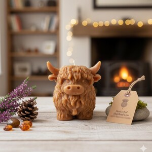 May include: A brown candle shaped like a Highland cow, with a wick on top. The candle has detailed fur and horns. A tag reads "Handcrafted Highland Cow". The candle sits on a wooden surface with a blurred background.
