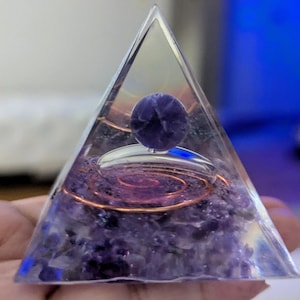 May include: A clear glass pyramid with a purple amethyst crystal at the top. The pyramid is filled with amethyst crystals and a copper wire coil.