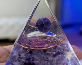 May include: A clear glass pyramid with a purple amethyst crystal at the top. The pyramid is filled with amethyst crystals and a copper wire coil.