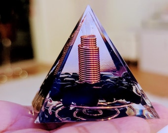 May include: A clear glass pyramid with a copper wire coil inside. The pyramid is filled with black stones and is sitting on a hand.