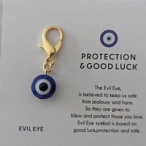 May include: A gold charm with a blue evil eye charm attached to a gold lobster clasp. The charm is on a white card with the text "EVIL EYE", "PROTECTION & GOOD LUCK", and a description of the evil eye charm.
