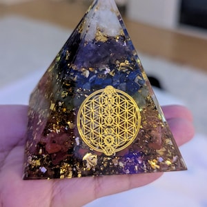 May include: A multi-colored crystal pyramid with gold flecks and a gold flower of life symbol in the center. The pyramid is made of clear resin and contains various crystals, including amethyst, rose quartz, and clear quartz.