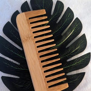 May include: A natural wooden comb with a rectangular shape and fine teeth. The comb is made of light brown bamboo and is resting on a dark green leaf.