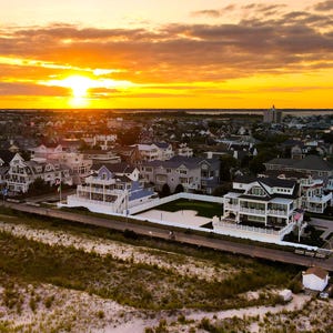 May include: An aerial view of a residential neighborhood with houses in a variety of styles. The houses are all white and have a variety of architectural details. The houses are set on a grassy lot with a sandy beach in the foreground. The sky is a vibrant orange and yellow, indicating a sunset.