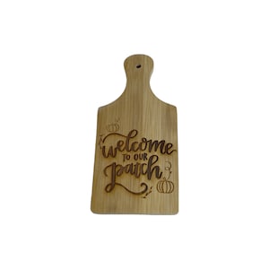 May include: A light brown wooden cutting board with a handle and a hole for hanging. The words "Welcome to our Patch" are laser-cut into the wood, along with pumpkin illustrations. The board has a rectangular shape.