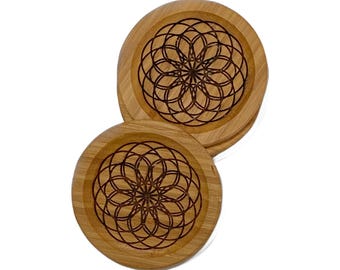 May include: Two round wooden coasters with a geometric floral design etched into the surface. The coasters are made of light-colored wood and have a dark brown, intricate pattern in the center. The coasters are approximately 4 inches in diameter.