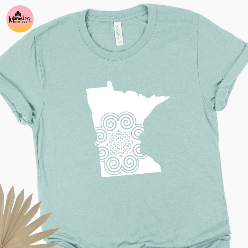 May include: A light blue t-shirt with a white outline of the state of Minnesota. The outline includes a white mandala design in the center.