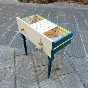 Könnte beinhalten: Eine kleine, handbemalte Holzschublade mit türkisfarbenen und cremefarbenen Akzenten. Die Schublade hat einen goldfarbenen Griff und wird von vier gedrechselten Beinen getragen, zwei türkisfarbenen und zwei cremefarbenen. Der Innenraum hat eine gemusterte Tapete.