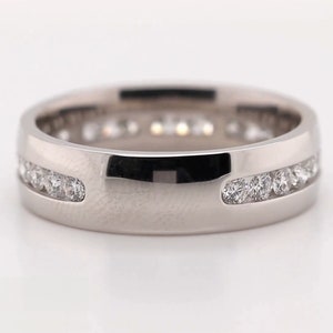May include: A silver-colored ring with a channel setting of small, clear gemstones. The ring has a flat, wide band with a polished finish. The gemstones are set in a continuous row around the ring. The ring is displayed on a white surface.