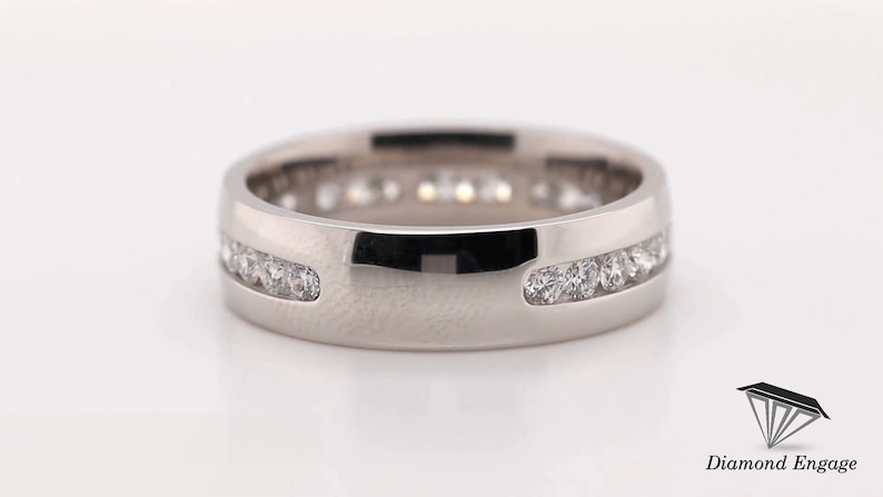May include: A silver-colored ring with a channel setting of small, clear gemstones. The ring has a flat, wide band with a polished finish. The gemstones are set in a continuous row around the ring. The ring is displayed on a white surface.