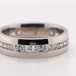 May include: A silver-colored ring with a channel setting of small, round, clear gemstones. The ring has a polished finish and a flat band design. The gemstones are set in two parallel rows around the ring.