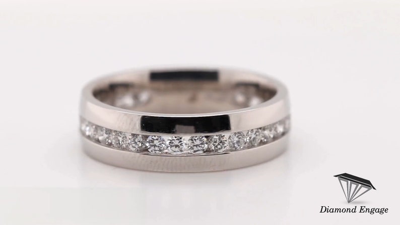 May include: A silver-colored ring with a channel setting of small, round, clear gemstones. The ring has a polished finish and a flat band design. The gemstones are set in two parallel rows around the ring.