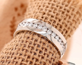 May include: A silver ring with a channel setting, showcasing a row of sparkling round-cut diamonds. The ring has a polished finish and a modern design, perfect for a special occasion. The ring is displayed on a textured, woven fabric.