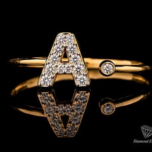 Peut inclure: Une bague ouverte en or ornée d'une lettre A majuscule incrustée de petites pierres claires et d'une pierre ronde à l'autre extrémité de l'anneau. La bague est présentée sur un fond noir avec le texte Diamond Engage dans le coin.