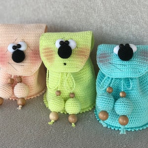 Op de afbeelding: Drie gehaakte pluche rugzakken in roze, groen en blauw met zwarte ogen en witte accenten. De rugzakken hebben een trekkoord sluiting en houten kralen.