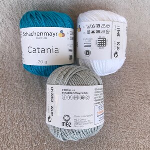 Puede incluir: Tres ovillos de lana en turquesa, gris y blanco. La lana est&aacute; etiquetada como "Schachenmayr Catania" y pesa 20 gramos. La lana est&aacute; hecha en Hungr&iacute;a para MEZ GmbH.
