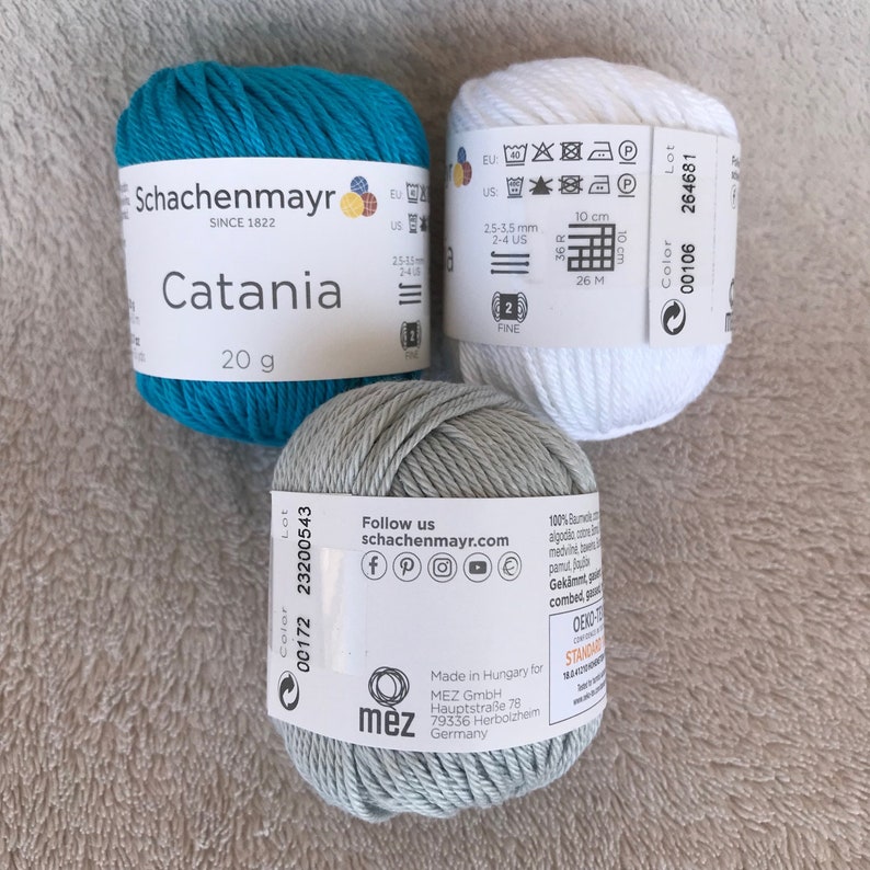 Puede incluir: Tres ovillos de lana en turquesa, gris y blanco. La lana est&aacute; etiquetada como "Schachenmayr Catania" y pesa 20 gramos. La lana est&aacute; hecha en Hungr&iacute;a para MEZ GmbH.
