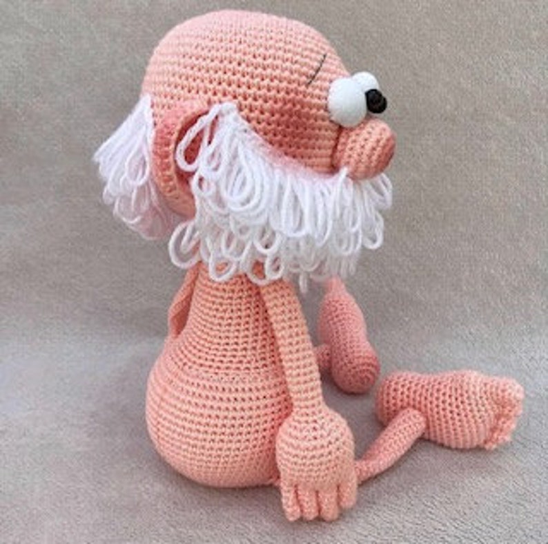 Puede incluir: Un juguete de peluche tejido a crochet con forma de persona con cuerpo rosa, barba blanca y ojos grandes. El juguete est&aacute; sentado con las piernas cruzadas.