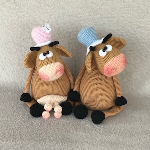 Puede incluir: Dos muñecas de vaca de ganchillo. Una vaca lleva un sombrero de copa rosa con una mariposa blanca, la otra un sombrero de copa azul. Ambas tienen cuerpos marrones, narices rosas y pezuñas negras. Están sentadas una al lado de la otra.