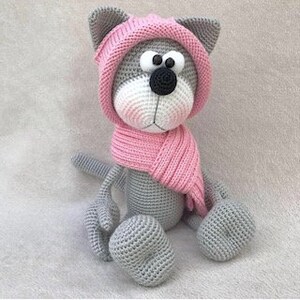Puede incluir: Gato de crochet gris con un gorro y una bufanda rosas. El gato est&aacute; sentado con las piernas cruzadas y tiene ojos grandes y negros.