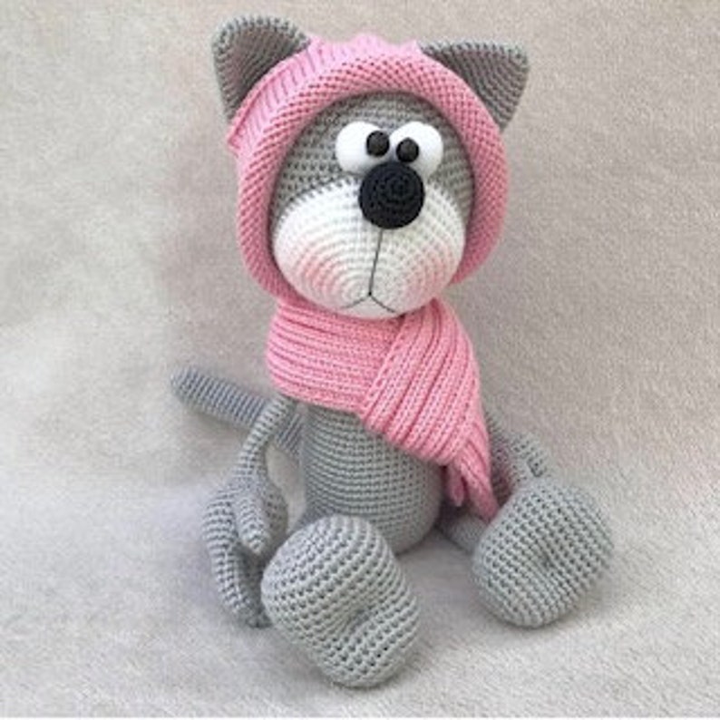 Puede incluir: Gato de crochet gris con un gorro y una bufanda rosas. El gato est&aacute; sentado con las piernas cruzadas y tiene ojos grandes y negros.