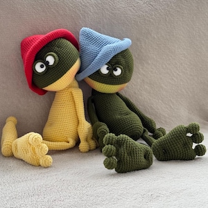 May include: Two handmade crochet frog dolls. One frog is yellow with a red hat, the other is green with a blue hat. Both have large, white-eyed faces and are sitting with their legs outstretched. The dolls are made of yarn.