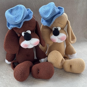 May include: Two handmade crochet dog plushies. One is brown, the other tan, both wearing blue hats. Each has a black nose, white muzzle with pink cheeks, and large, expressive eyes. Detached legs are next to each dog.