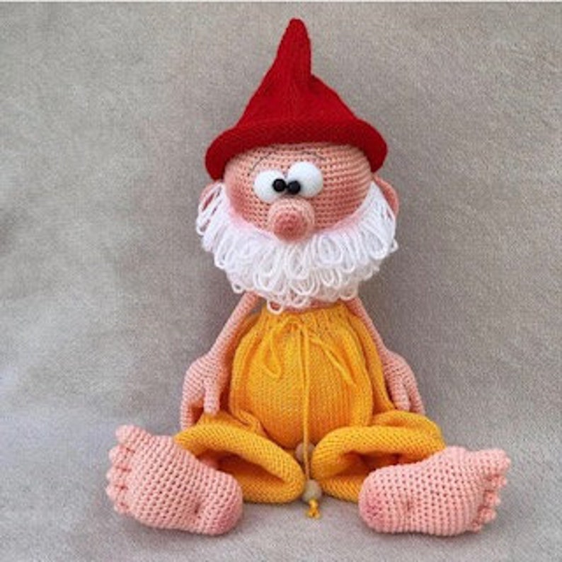 Puede incluir: Un mu&ntilde;eco de gnomo de crochet con un gorro rojo, barba blanca y pantalones amarillos. El gnomo est&aacute; sentado con las piernas cruzadas y tiene una nariz grande y redonda.