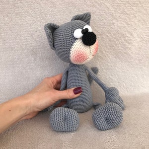 Puede incluir: Un juguete de crochet de gato gris con grandes ojos blancos, una nariz negra y mejillas rosadas. El gato est&aacute; sentado sobre una superficie gris suave.