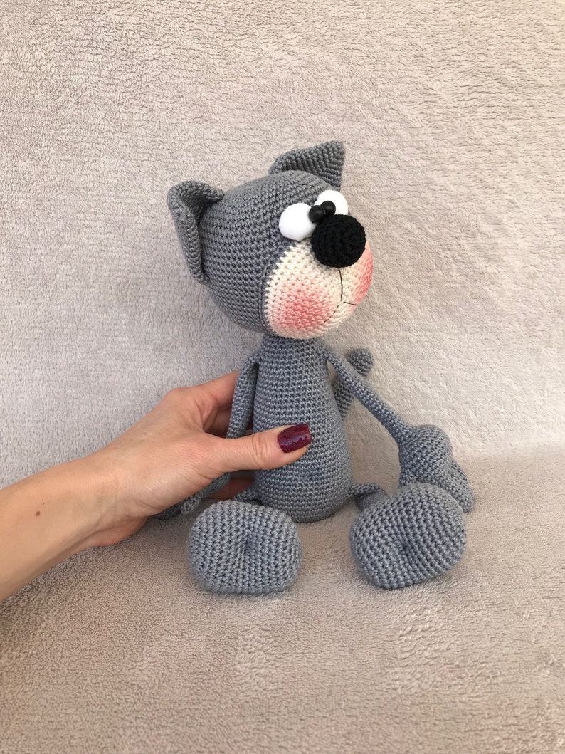 Puede incluir: Un juguete de crochet de gato gris con grandes ojos blancos, una nariz negra y mejillas rosadas. El gato est&aacute; sentado sobre una superficie gris suave.