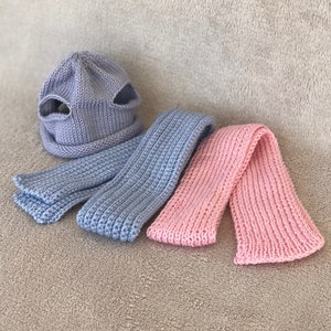 Puede incluir: Un conjunto de dos bufandas de punto y un gorro. El gorro es azul claro y tiene dos agujeros en la parte superior. Una bufanda es azul claro y la otra es rosa.