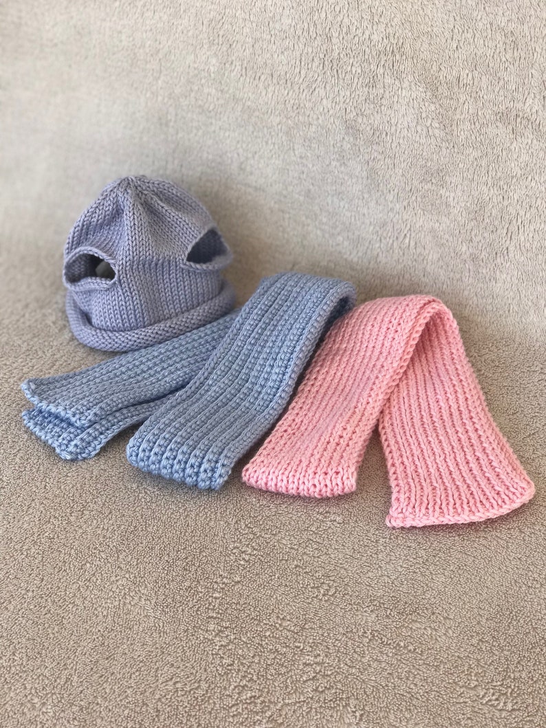 Puede incluir: Un conjunto de dos bufandas de punto y un gorro. El gorro es azul claro y tiene dos agujeros en la parte superior. Una bufanda es azul claro y la otra es rosa.