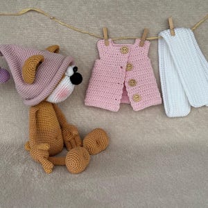 May include: A crocheted stuffed animal wearing a pink hat with a brown body and a white face. The animal is sitting on a light brown surface. There is a pink crocheted vest and a white crocheted scarf hanging in the background.