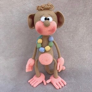 Puede incluir: Un juguete de mono de crochet con pelaje marr&oacute;n, manos y pies rosas y un collar de cuentas de colores. El mono tiene ojos grandes y negros y una nariz rosa.