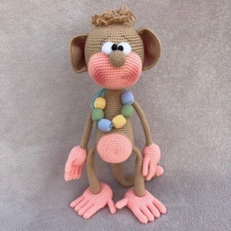 Puede incluir: Un juguete de mono de crochet con pelaje marr&oacute;n, manos y pies rosas y un collar de cuentas de colores. El mono tiene ojos grandes y negros y una nariz rosa.