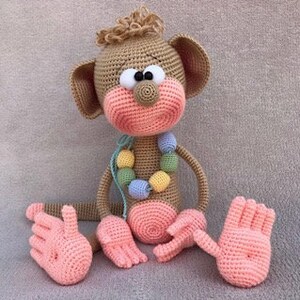 Puede incluir: Un juguete de mono de crochet con pelaje marr&oacute;n, manos y pies rosas y un collar de cuentas de colores.