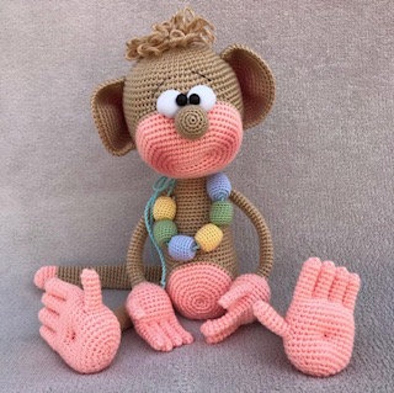 Puede incluir: Un juguete de mono de crochet con pelaje marr&oacute;n, manos y pies rosas y un collar de cuentas de colores.