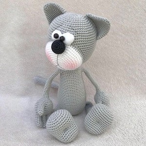 Puede incluir: Un juguete de crochet de gato gris con rasgos faciales blancos, ojos negros y nariz rosa. El gato est&aacute; sentado con las piernas extendidas.