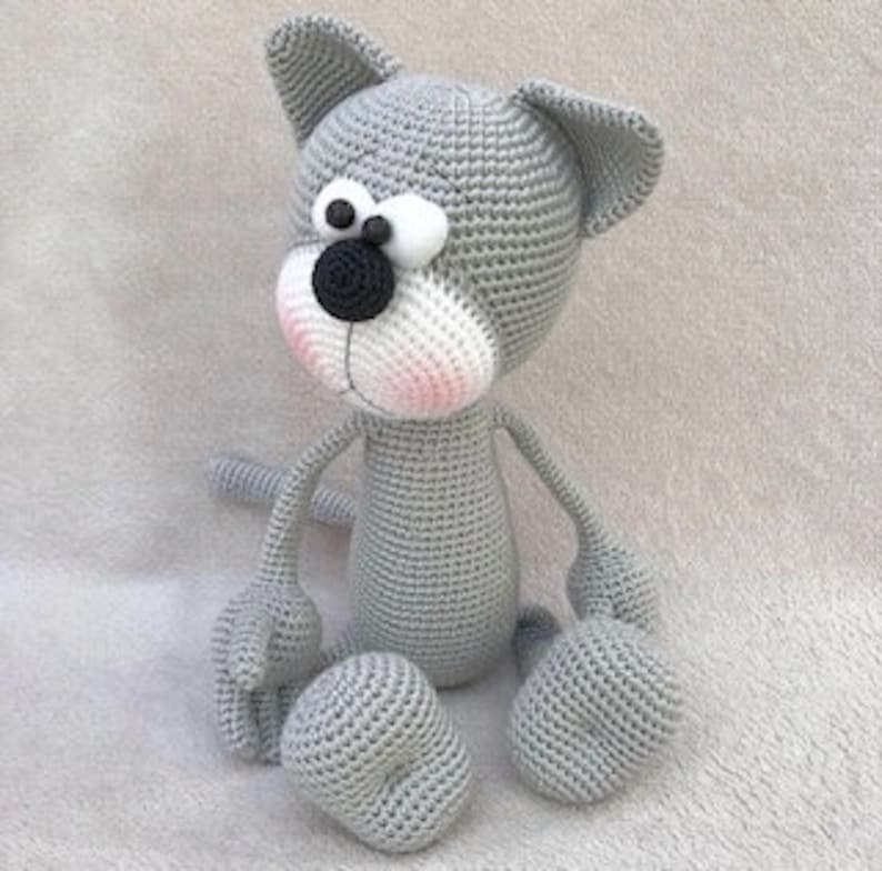 Puede incluir: Un juguete de crochet de gato gris con rasgos faciales blancos, ojos negros y nariz rosa. El gato est&aacute; sentado con las piernas extendidas.