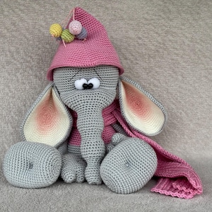 May include: A grey crocheted elephant wearing a pink crocheted hat with colourful pom-poms and a pink scarf.