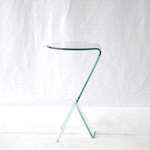 Könnte beinhalten: Ein Beistelltisch aus klarem Glas, der aus einem einzigen, durchgehend gebogenen Stück Glas besteht und eine runde Tischplatte sowie einen Z-förmigen Fuß bildet. Die Struktur ist transparent mit grünlich getönten Kanten.