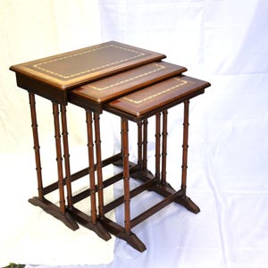 May include: A set of three nested tables with dark brown wooden frames and bamboo-style legs. Each rectangular tabletop features a smooth, dark brown surface with a decorative gold-coloured border inlay around the perimeter of the top edge.